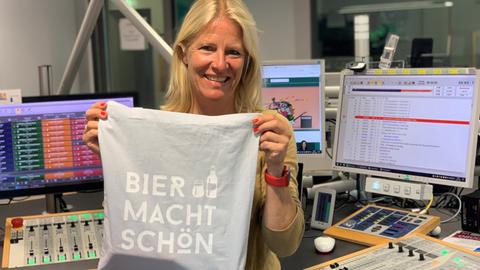 Britta Lohmann steht im Studio und hält einen Jutebeutel mit der Aufschrift: Bier macht schön in die Kamera.