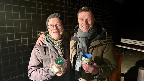 Zwei Männer stehen im Dunkeln draußen. Sie tragen Winterkleidung. Der eine hält eine Tasse in der Hand, der andere ein Glas mit Süßigkeiten.