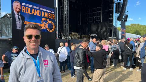 hr4-Reporter Peter Küster steht vor der Bühne in Willingen. Er trägt eine Sonnenbrille und eine graue Sweatshirtjacke mit hr4-Logo.