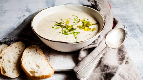 Ein Teller mit Käsesuppe steht auf einem Tisch, daneben liegen silberne Löffel und ein paar Scheiben Baguette.