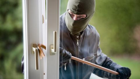 Ein maskierter Einbrecher macht sich mit einem Brecheisen an einem Fenster zu schaffen.